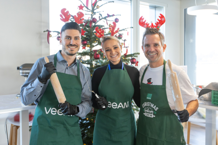 Three people in green aprons stand in front of a Christmas tree.