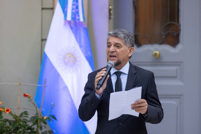 A man in a suit speaks into a microphone in front of an Argentinian flag.