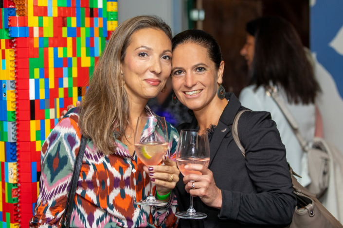 Two women pose for a photo in front of a Lego wall.