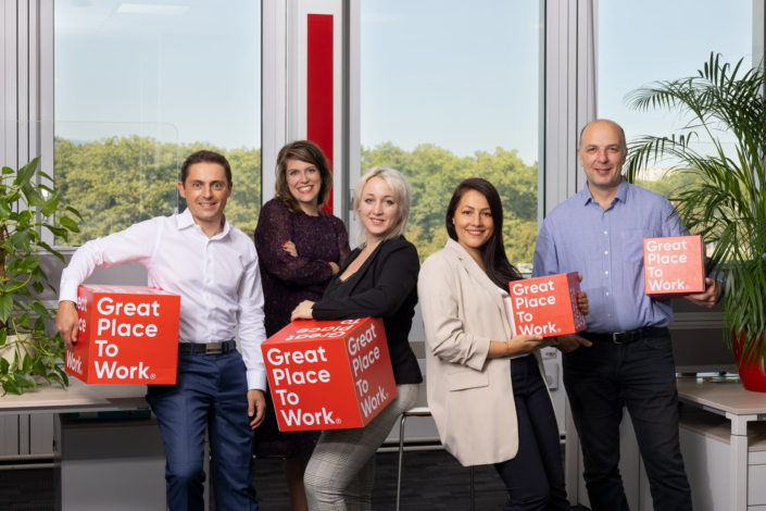Eine Gruppe von Menschen posiert vor einem roten Kasten mit der Aufschrift „Great Place to Work“.
