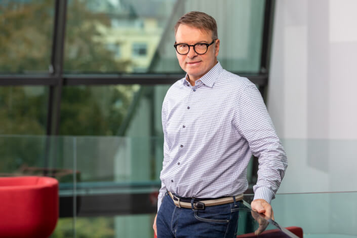 A man with glasses stands in front of a glass wall.