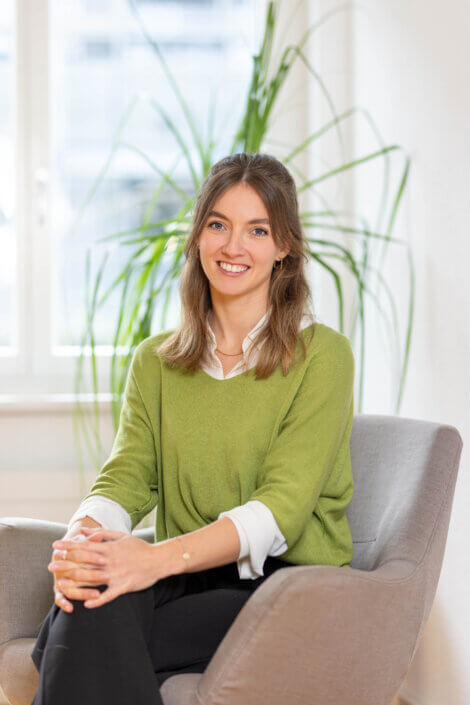 A person with long hair, wearing a green sweater and a white shirt, sits smiling on a gray chair. A plant stands next to a window in the background.