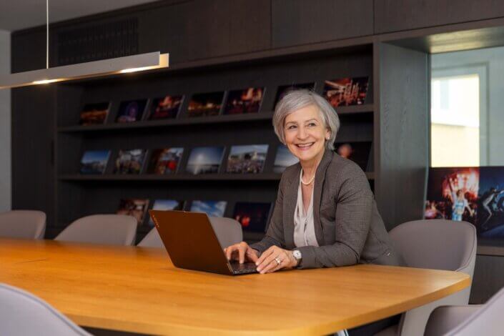 An older woman with short gray hair smiles as she works on a laptop at a wooden conference table. She is in a modern office, with shelves with various framed pictures in the background.