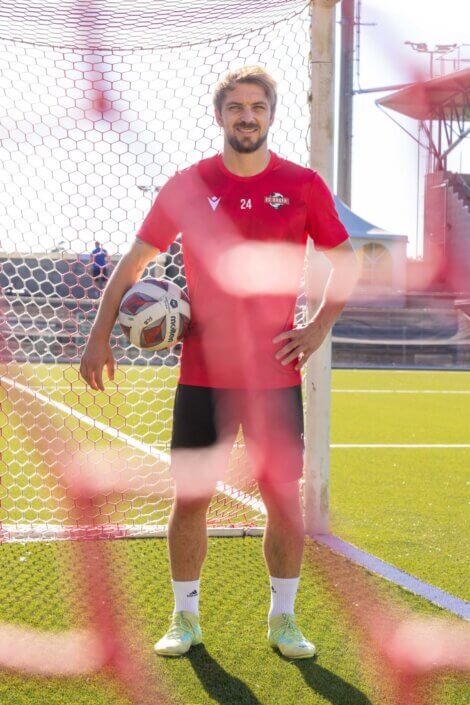 A soccer player in a red jersey and black shorts stands in a goal on a sunny day and holds a soccer ball under his arm.