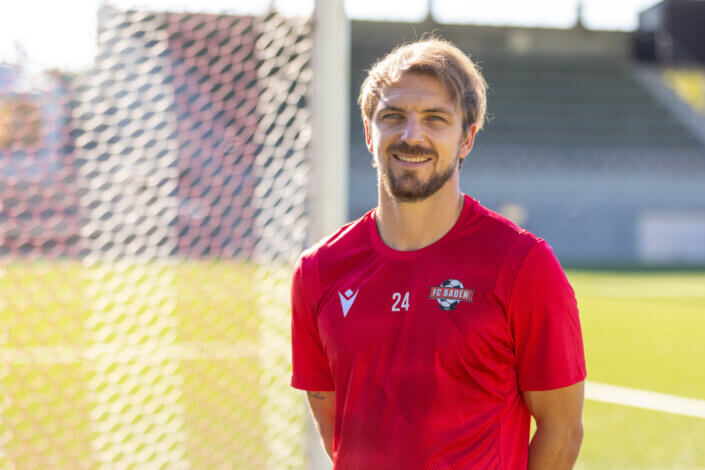 A soccer player in a red jersey with the number 24 stands on a field. A goal and empty stands can be seen in the background.