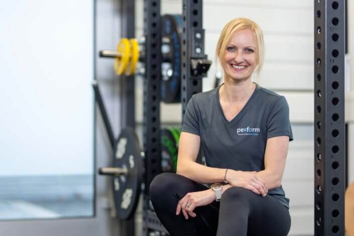 A woman in a gray "Perform" shirt sits smiling next to fitness equipment, weights can be seen in the background.