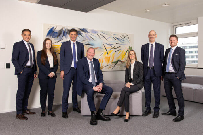 A group of six men and one woman in business attire pose in an office with abstract artwork in the background.