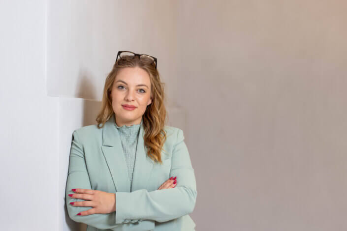A woman with long hair, glasses on her head and a light green blazer stands with her arms crossed, leaning against a white wall.