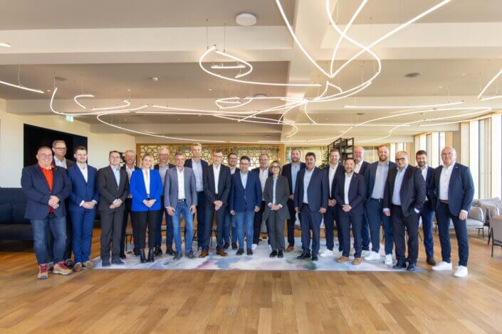 A group of people in business attire stand in a modern, well-lit room with a decorative ceiling design.