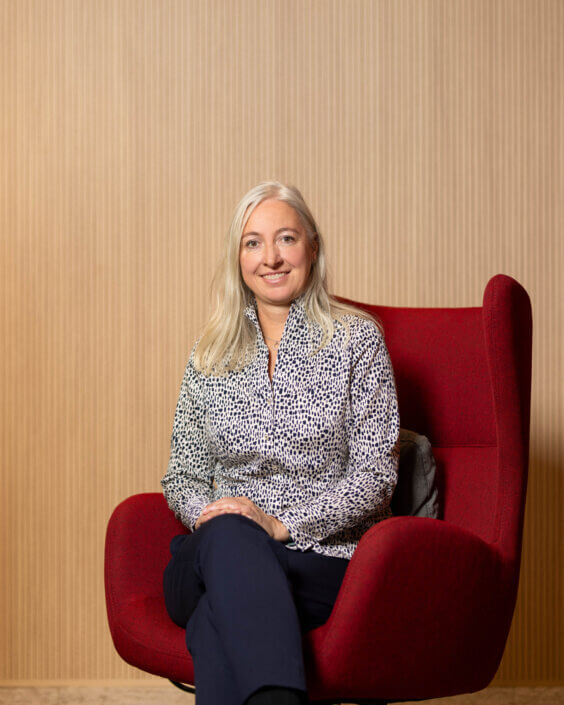 A woman with long blonde hair, a patterned blouse and dark pants sits on a red chair in front of a light-colored wooden background. She is smiling and has her hands in her lap.