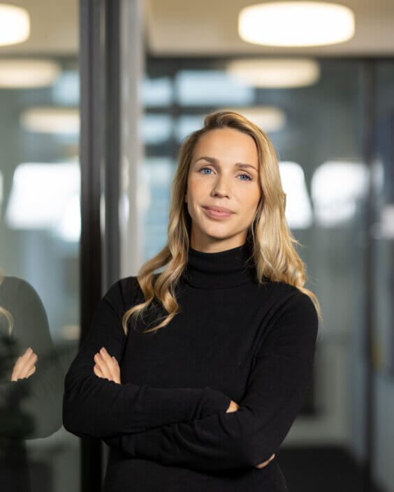 A woman in a black turtleneck sweater stands with her arms folded against a glass wall in a well-lit room.