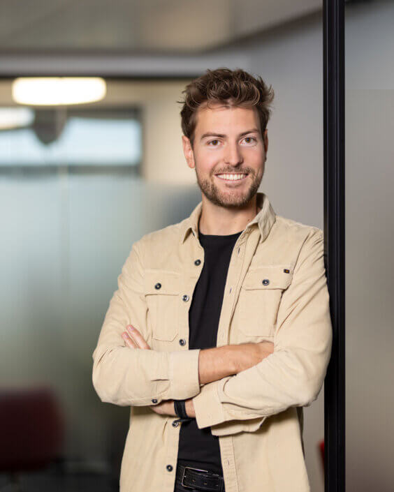 A person with short hair and a beard, wearing a beige shirt and a black undershirt, stands with folded arms in an office environment.