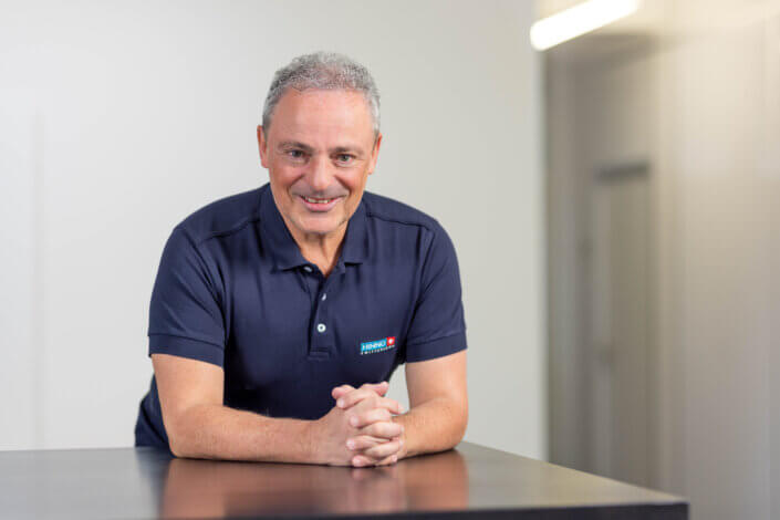 Smiling man with short gray hair in a blue polo shirt, leaning against a dark table, indoors against a neutral background.