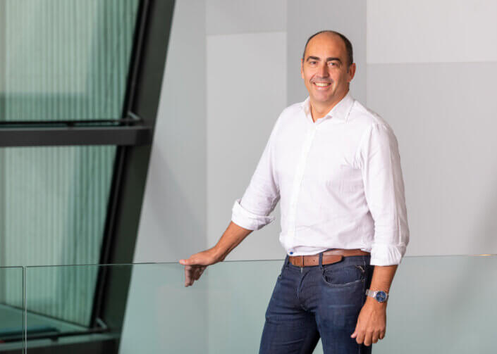 A man in a white shirt and jeans stands smiling in the house and rests one hand on a glass railing.