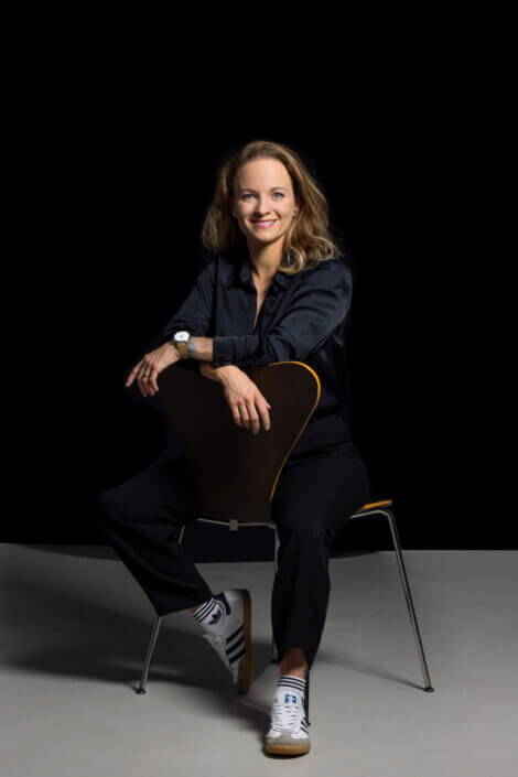 A woman in a dark outfit and white sneakers sits smiling on a chair in front of a black background.