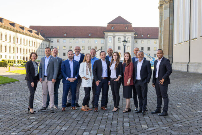 A group of 16 people, both men and women, stand together outside on a cobblestone path with a large building in the background.