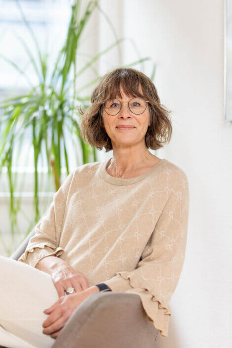 Middle-aged woman with glasses is sitting inside, holding a book in her hand. She has shoulder-length brown hair and is wearing a beige sweater. A leafy plant can be seen in the background.