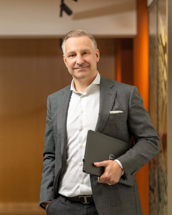 A person in a gray suit and white shirt stands inside holding a closed Dell laptop in front of a blurred background.