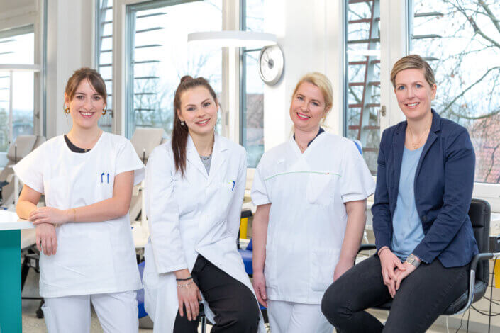 Four women, three in white uniforms and one in a blue blazer, smile in a professional interior.