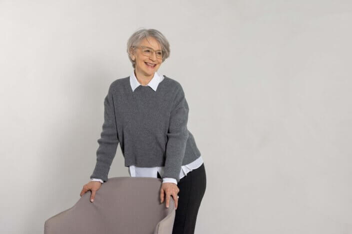 An older woman with short gray hair and glasses stands behind a chair, smiling. She is wearing a gray sweater over a white shirt and black trousers.