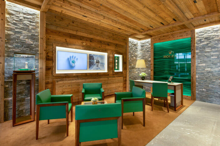 Luxurious interior of a watch store with wood and stone decor, green chairs, showcases and Rolex logo on the walls.
