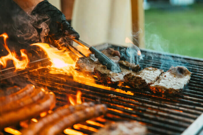 Grill mit Würstchen und Fleisch, das über offenem Feuer gegart wird. Eine behandschuhte Hand wendet das Fleisch mit einer Zange.