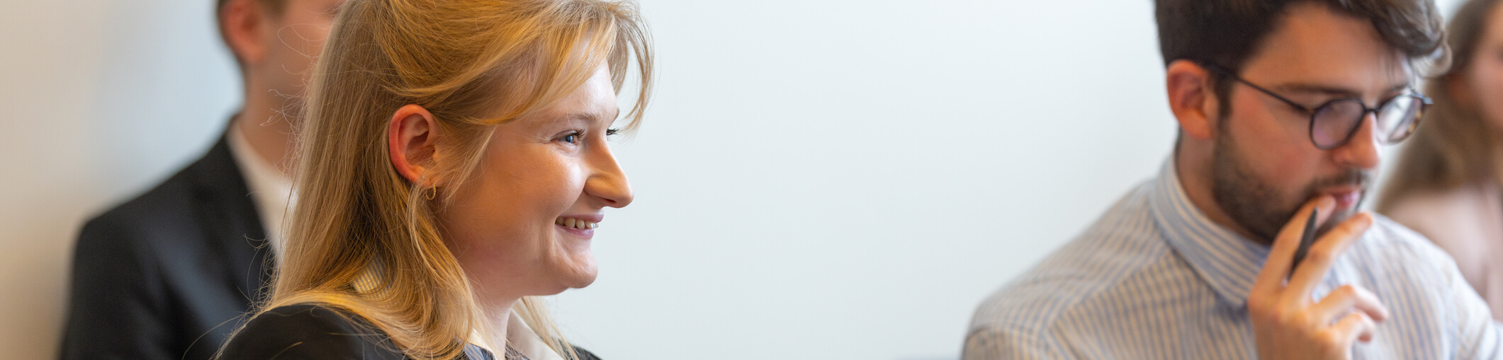 Eine Frau und ein Mann sitzen in einem Besprechungsraum. Die Frau lächelt, der Mann hält die Hand vor den Mund. Er trägt eine Brille und ein gestreiftes Hemd. Im Hintergrund ist eine weitere verschwommene Figur zu sehen.