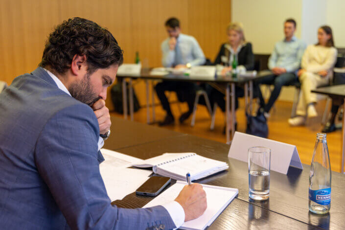 Ein Mann schreibt an einem Konferenztisch mit Dokumenten und einem Telefon. Zwei Männer und zwei Frauen sitzen an einem weiteren Tisch im Hintergrund. Auf beiden Tischen stehen Wasserflaschen.
