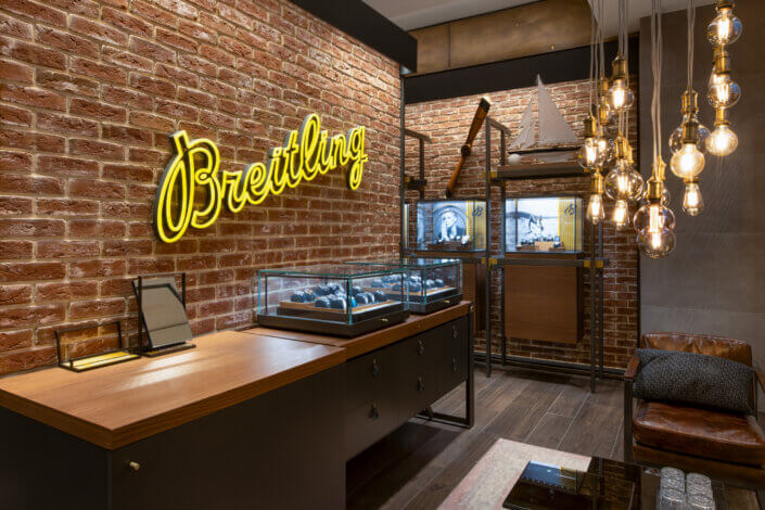 A stylish showroom with a neon-colored "Breitling" sign. Watches in glass showcases are presented in front of a brick wall. Hanging light bulbs illuminate the room.