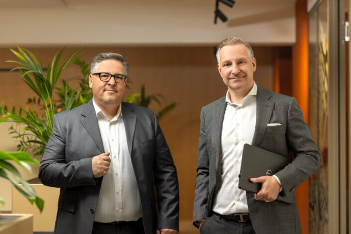 Two men in suits are standing in an office corridor. One is holding a laptop, the other is straightening his jacket. They are standing in front of potted plants and wooden walls.