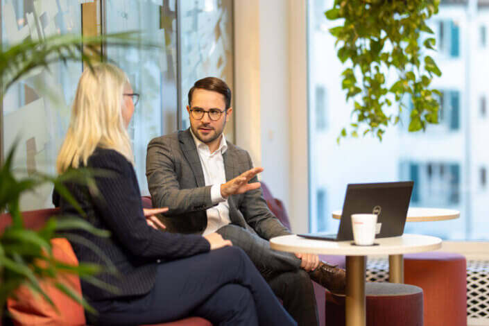 Two people in business attire are talking in an office. Between them are a laptop and a coffee cup on a small table. A large window and a hanging plant can be seen in the background.