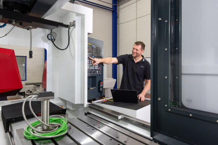 A man operates a CNC machine in a workshop. He points to the control panel while standing next to a laptop on a table. A green cable reel can be seen nearby.