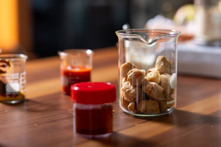 Cup filled with brown round snacks on a wooden table, next to a small glass with a lid and another cup with red liquid.