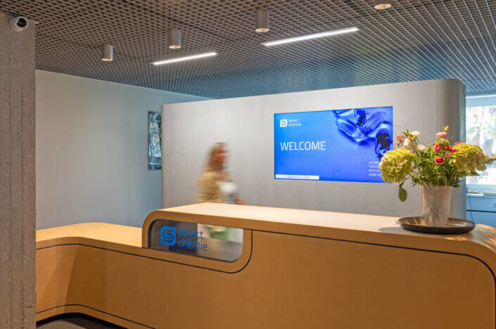 Reception area with a wooden desk and a digital billboard with the message "Welcome". A blurred person stands behind the desk. There are also flowers in a vase.