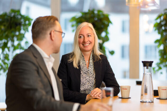 A woman in a business suit sits smiling with a man at a table in an office. There are plants and coffee cups on the table.