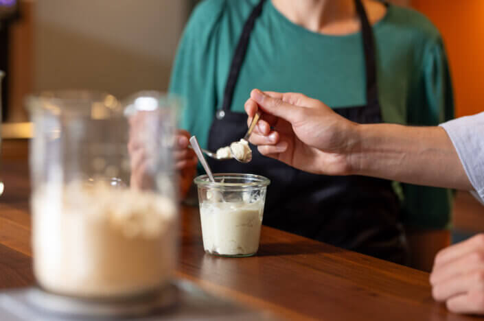 A person in an apron holds a jar of creamy mixture on a wooden table while another person spoons some of it out.