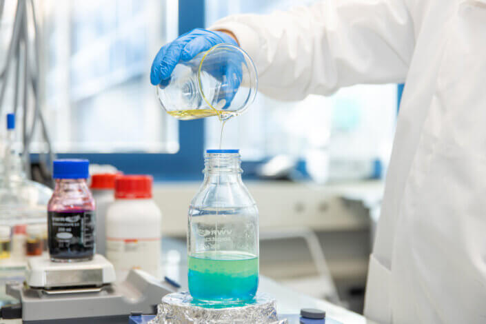 In a laboratory environment, a person in a lab coat and blue gloves pours a yellow liquid from a beaker into a transparent bottle containing a green liquid.