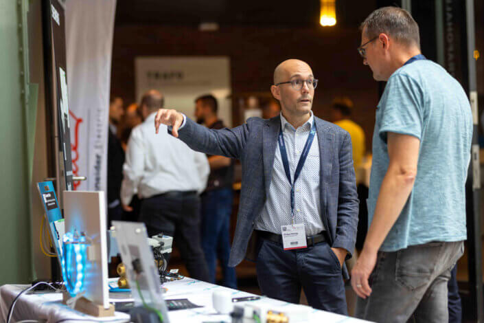 Zwei Männer unterhalten sich an einem Stand einer Technologiekonferenz. Auf dem Tisch sind elektronische Geräte ausgestellt.