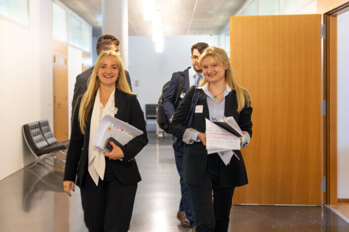 Zwei Frauen in Businesskleidung gehen mit Dokumenten einen Flur entlang. Drei weitere Personen folgen ihnen, alle in Berufskleidung. Im Hintergrund sind Türen und Sitzgelegenheiten zu sehen.