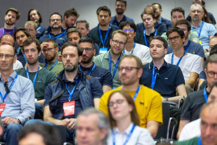 Bei einer Indoor-Veranstaltung hören die Zuschauer mit Lanyards aufmerksam zu. Das Publikum wirkt engagiert und vielfältig in Alter und Aussehen.