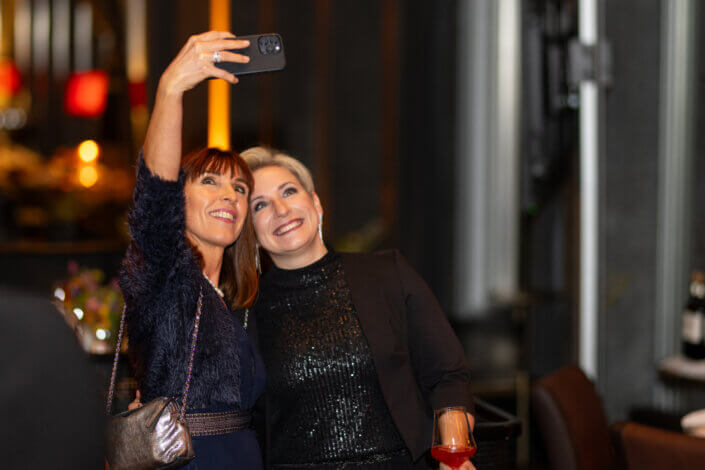 Zwei Personen stehen in einem warm beleuchteten Innenraum eng beieinander und machen ein Selfie. Eine hält mit der linken Hand ein Smartphone hoch, beide lächeln.