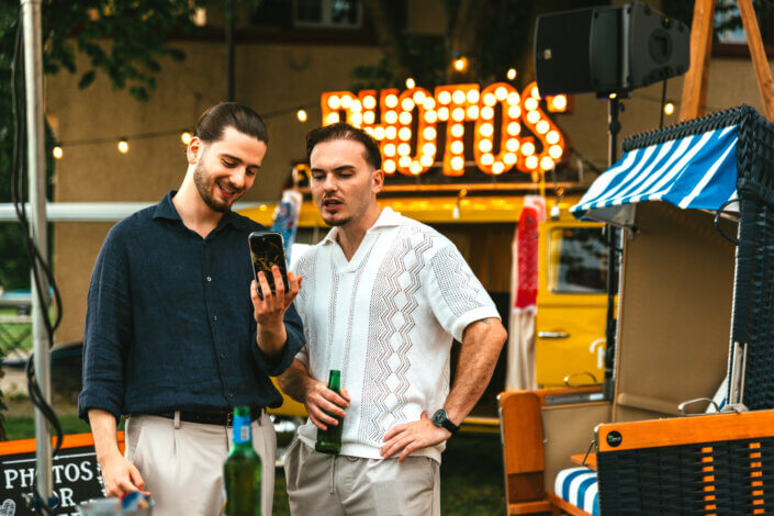 Zwei Personen stehen im Freien und schauen auf ein Telefon. Eine Person hält ein Bier in der Hand. Im Hintergrund sind ein leuchtendes „FOTOS“-Schild und ein gelbes Fahrzeug zu sehen.