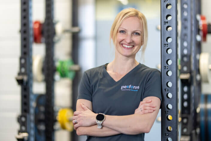 Smiling person in a fitness studio. She is wearing a gray T-shirt with the word "Perform" on it. She is standing in front of fitness equipment, including dumbbell racks.