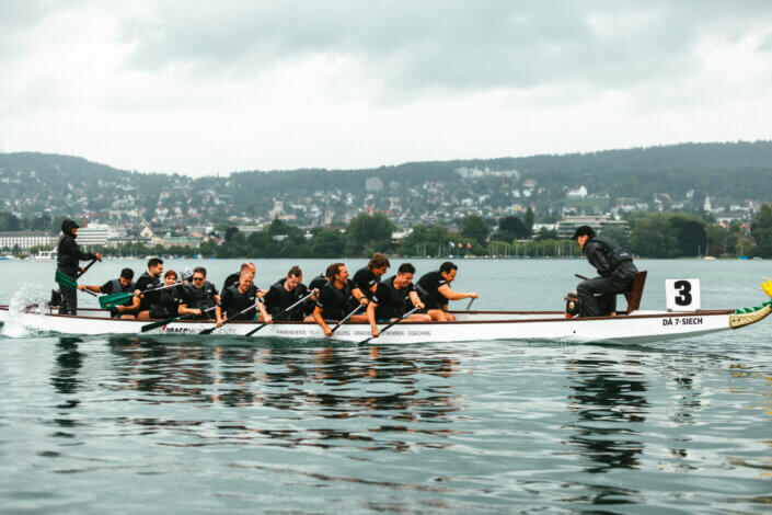 Eine Gruppe von Menschen rudert in einem Langboot mit der Aufschrift „3“ auf einem Gewässer, im Hintergrund ein bewölkter Himmel und ferne Hügel.