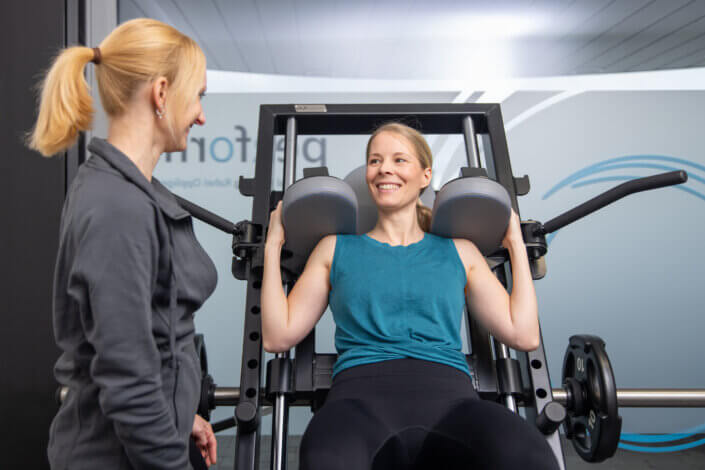 A woman in sportswear smilingly uses a leg press while another woman in a jacket stands next to her in a gym.