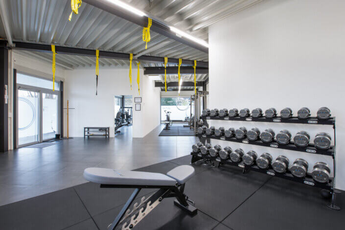 Spacious gym with a row of silver dumbbells on stands, a training bench and yellow resistance bands hanging from the ceiling.
