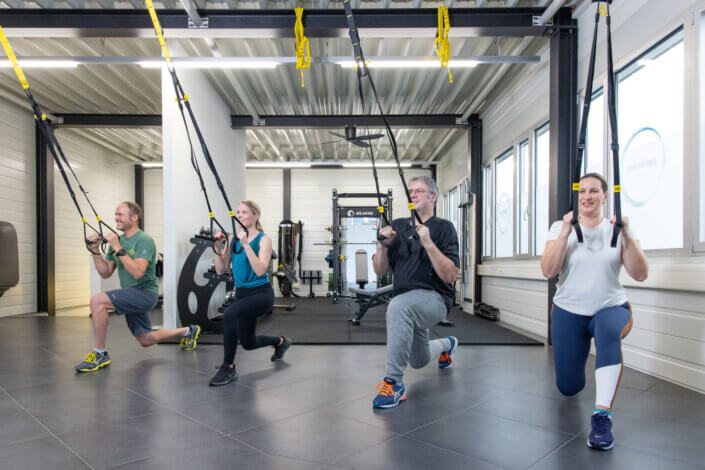 Four people are working out in a gym with sling straps and doing lunges. They are in a bright room with fitness equipment in the background.