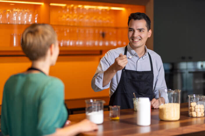 A man in a black apron smiles while holding a spoon and talking to a woman at a wooden counter with jars of ingredients.