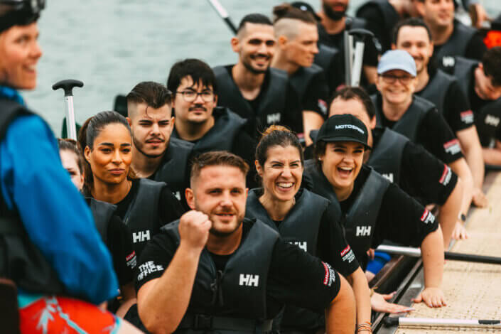 Eine Gruppe von Menschen mit Schwimmwesten sitzt auf einem Drachenboot im Wasser. Einige lächeln und halten Paddel in der Hand, bereit für eine Teamaktivität.
