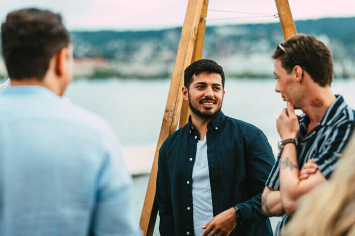Drei Personen unterhalten sich im Freien in der Nähe eines Gewässers. Ein Mann im dunklen Hemd spricht, während ein anderer im gestreiften Hemd aufmerksam zuhört. Eine dritte Person steht in der Nähe.